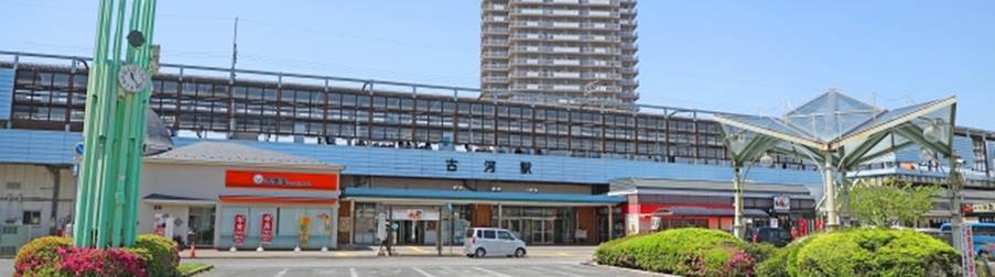 青空の下にある古河駅の駅舎と周辺の建物、時計塔と植え込みが見える風景