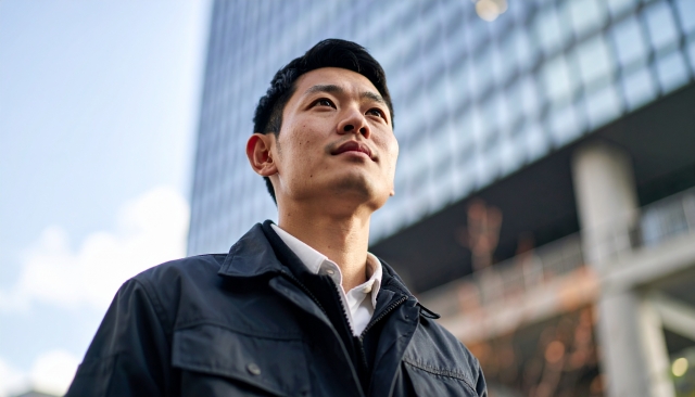 高層ビルの斜め上を見つめる若い作業服男性